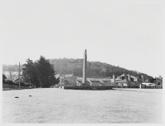 Black and white photo of Sueno's Stone, a tall carved cross slab, standing on grassy parkland with houses behind and a wooded hill behind that.