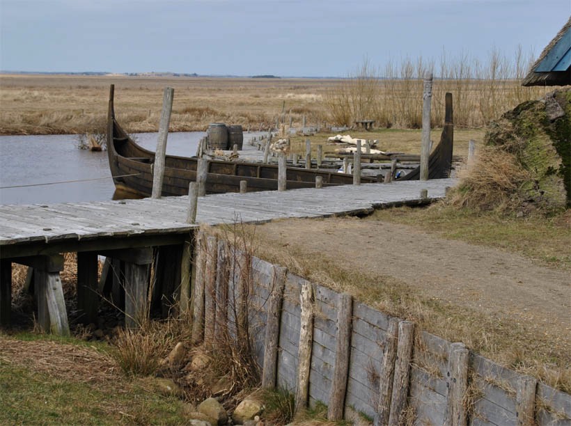 A reconstructed Viking trading harbour at Bork in Denmark. A viking longboat is moored by a wooden pontoon or jetty. Again this structure is flat, featuring planks laid across wooden trestles.