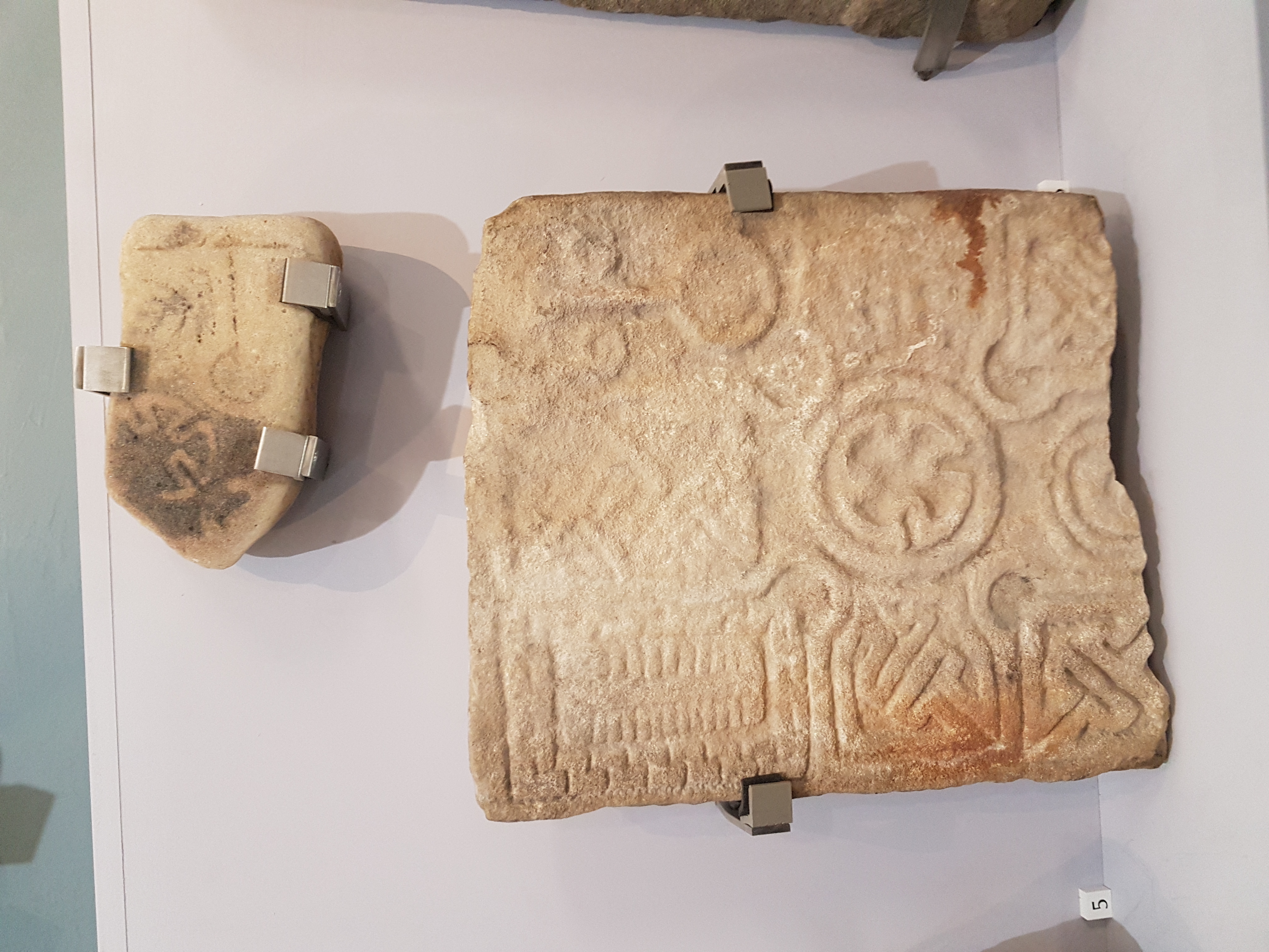 The Kinloss Stone and Drainie 32 displayed together in Elgin Museum. Drainie 32 is larger and has a cross head with interlace-filled arms and a Maltese cross-style roundel in the centre. Pictish mirror and comb symbols flank the top arm of the cross. There is key pattern decoration flanking the shaft of the cross. The Kinloss Stone is a smaller fragment,, with a similar Maltese cross-style roundel and more decoration that is quite hard to make out. (Accession Number ELGNM 2019.10)
